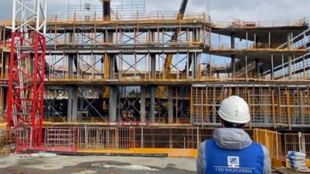 Operaio con casco bianco osserva un edificio in costruzione con struttura in cemento armato e ponteggi, presso un cantiere edile moderno.